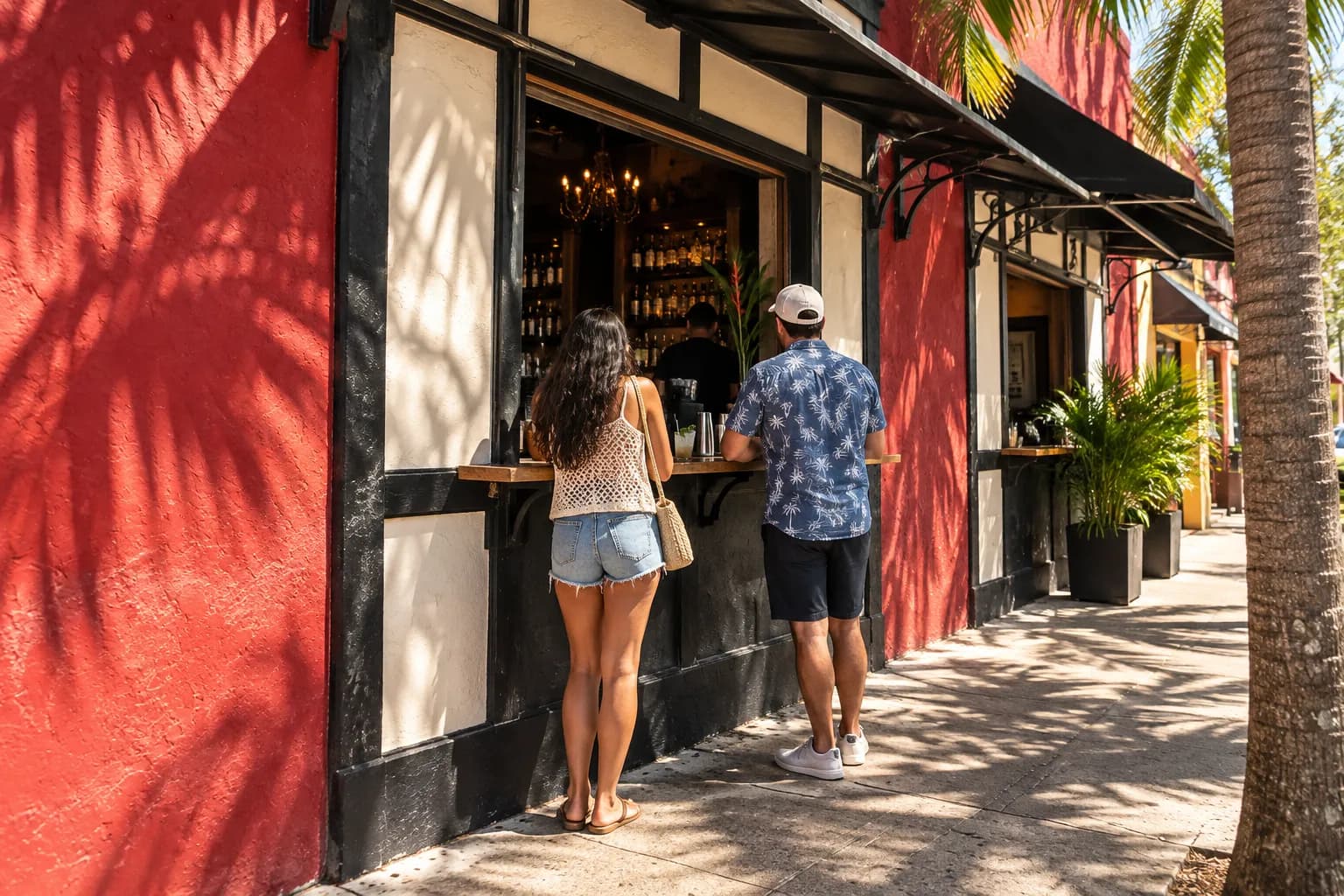 A small serving window cut into a painted wall with a narrow shelf beneath it, two people standing at it in mid-morning sun with palm shadows