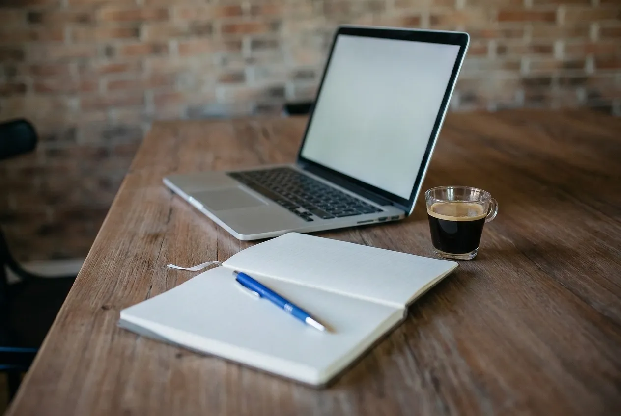 A laptop, notebook and coffee cup on a large shared wooden table by a window