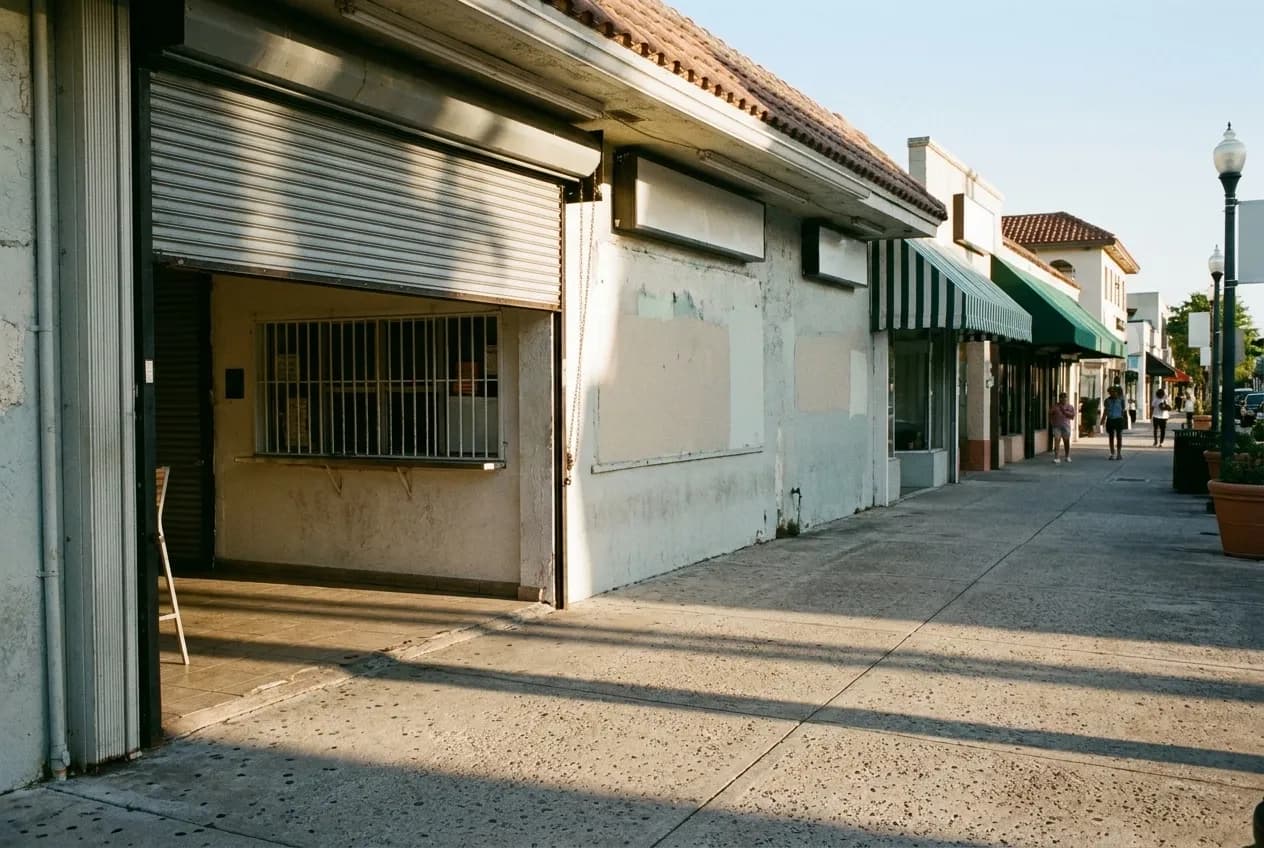 A low commercial street in early morning light with roller shutters half raised and a barred service window in a painted wall, long shadows across the pavement