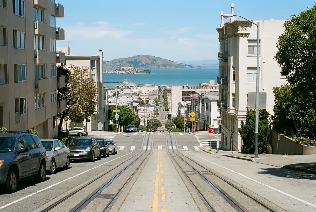 A steep San Francisco street with a cable car track and bay water in the distance