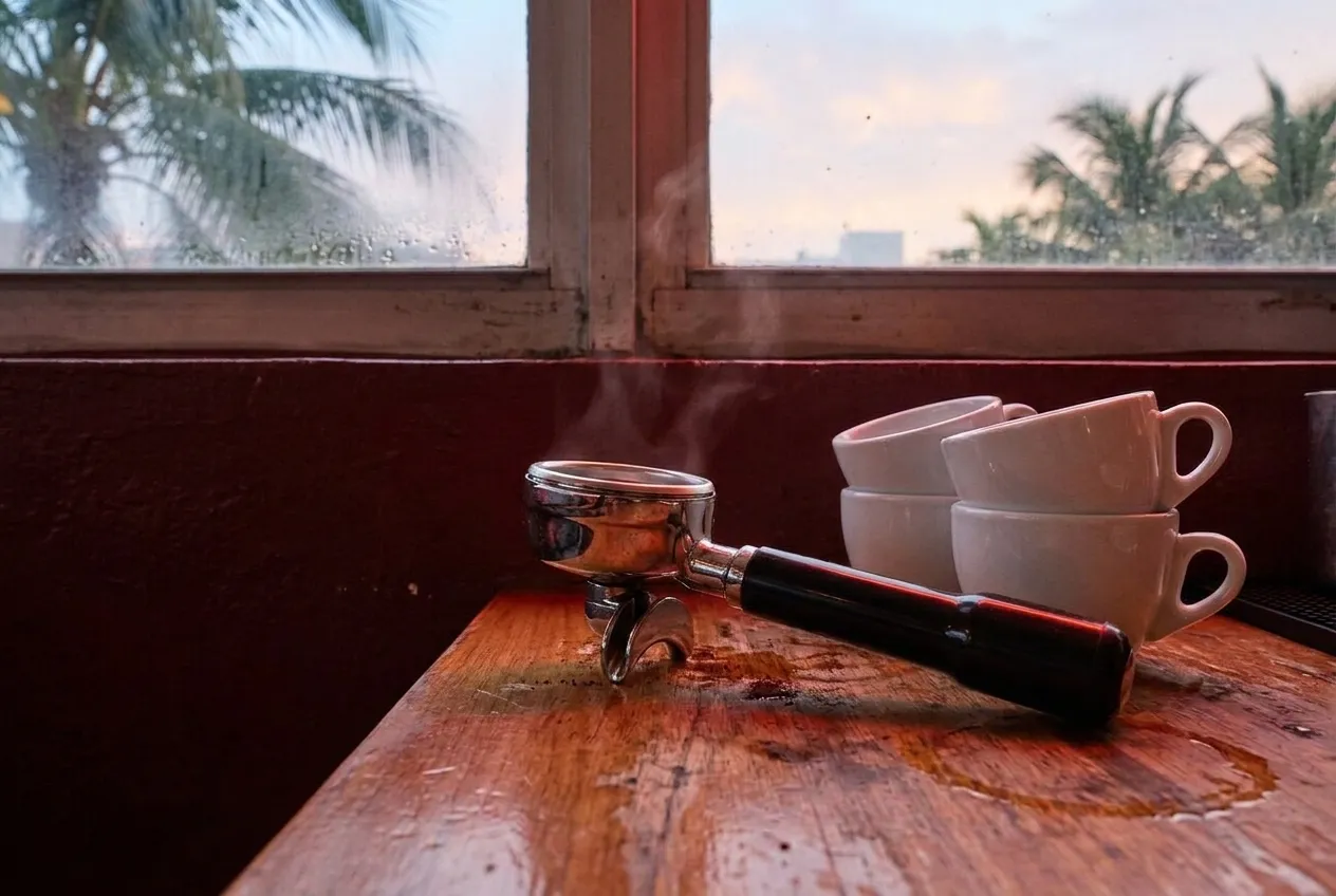 An espresso machine portafilter and tamped coffee grounds on a steel counter, with steam wands and a row of small cups in soft early light.