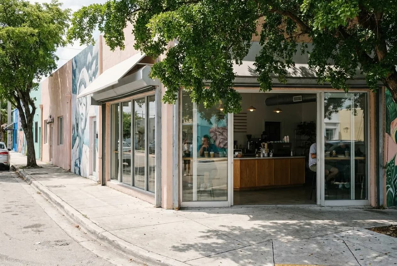 Sunlit Wynwood street corner with murals on low-rise buildings, dappled shade from tree canopy, and pedestrians walking past art-district storefronts in late morning light.