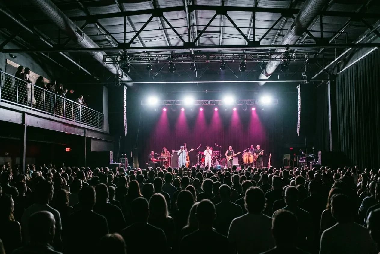 A wide view over a five-hundred-strong crowd toward a band lit in white and magenta in a modern Miami music room with a mezzanine rail above
