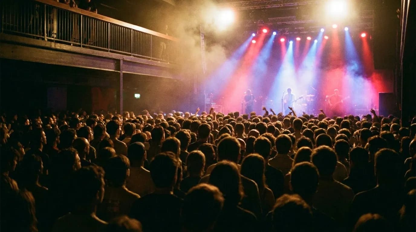 A packed standing floor at a mid-size music venue with a lit stage in the distance and coloured beams cutting through haze