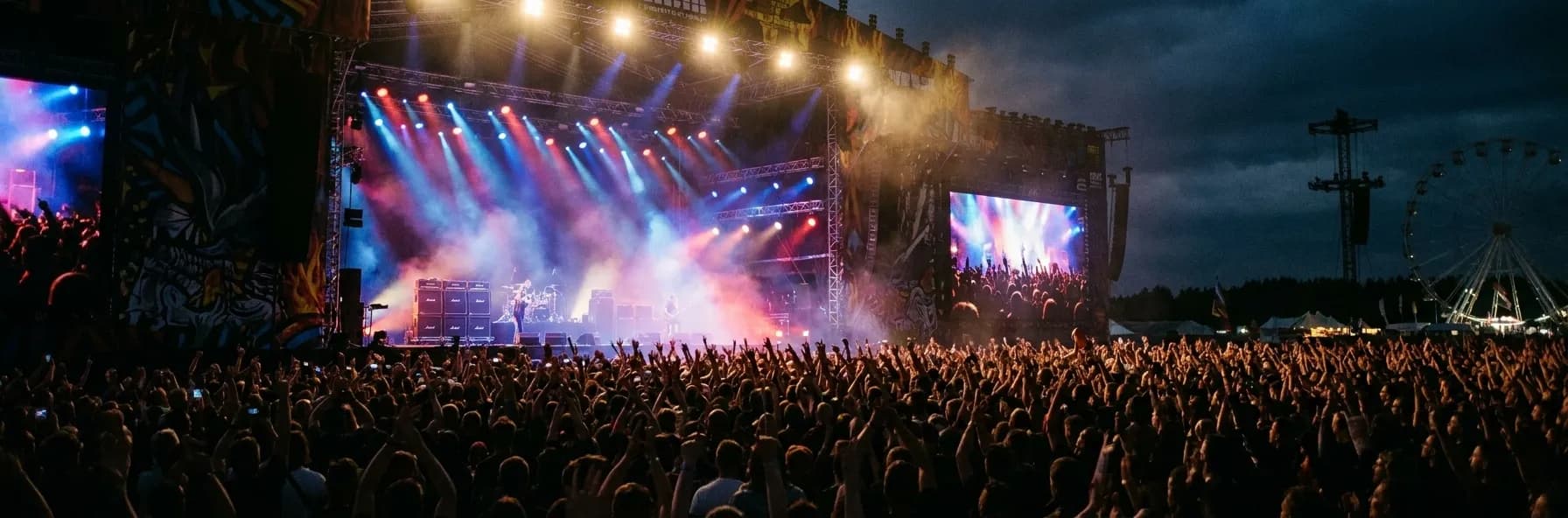 A concert stage seen from the middle of a standing crowd with coloured lights and raised hands in silhouette