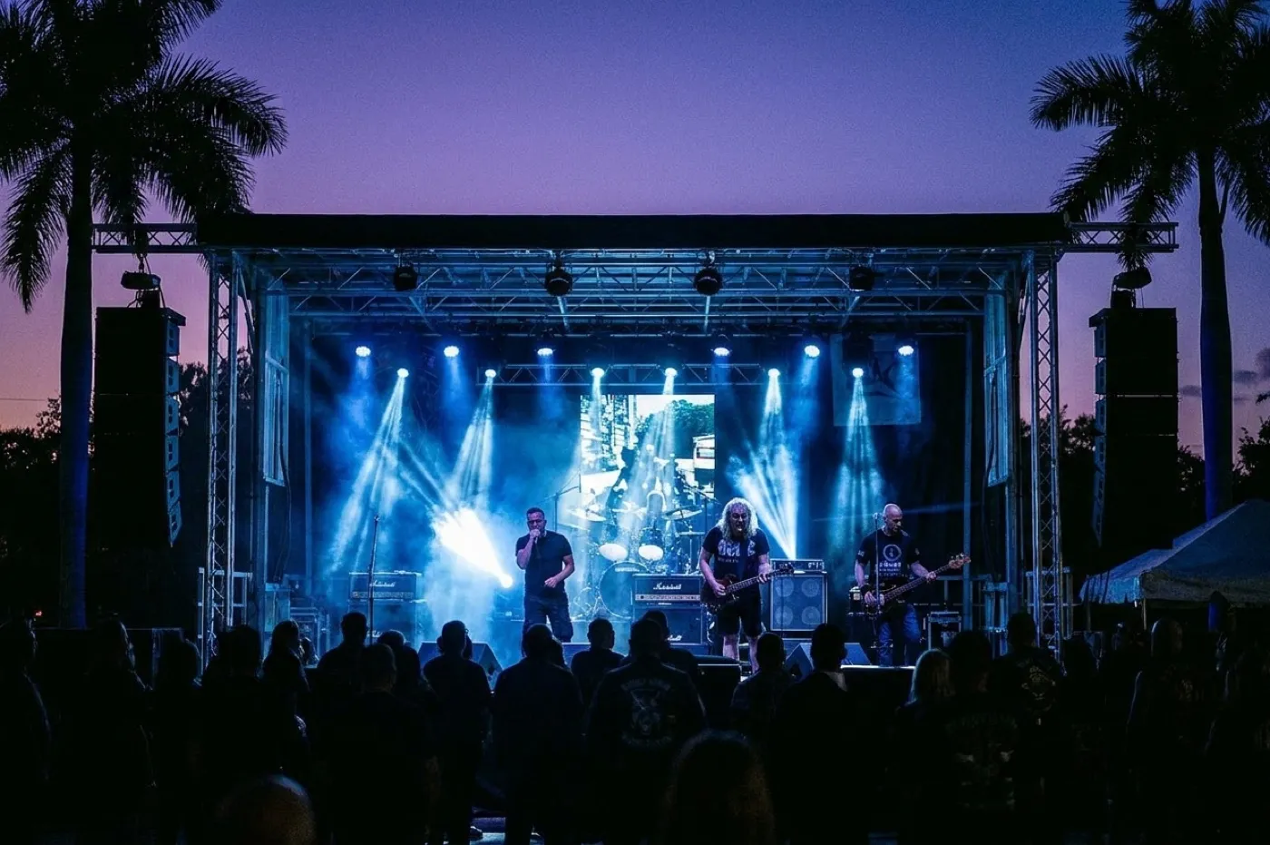 An outdoor concert stage at dusk with lighting rigs, a half-full crowd in silhouette and palms against a violet sky
