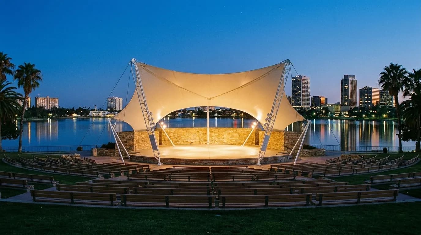 An outdoor amphitheatre stage at dusk seen from the lawn, empty seating in front and city towers rising behind the shell