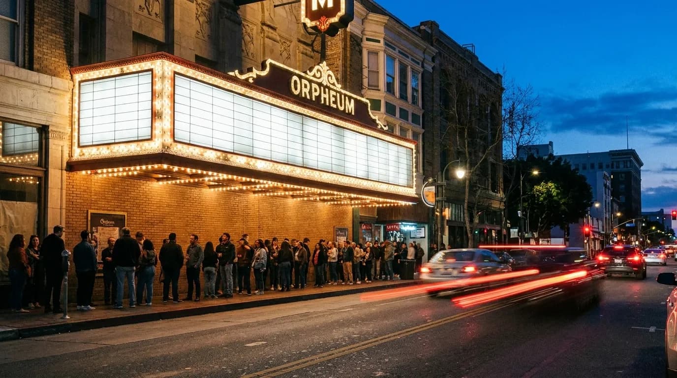 A queue outside a mid-size Los Angeles music venue at dusk, people waiting along a wall under a lit marquee with cars passing on the street