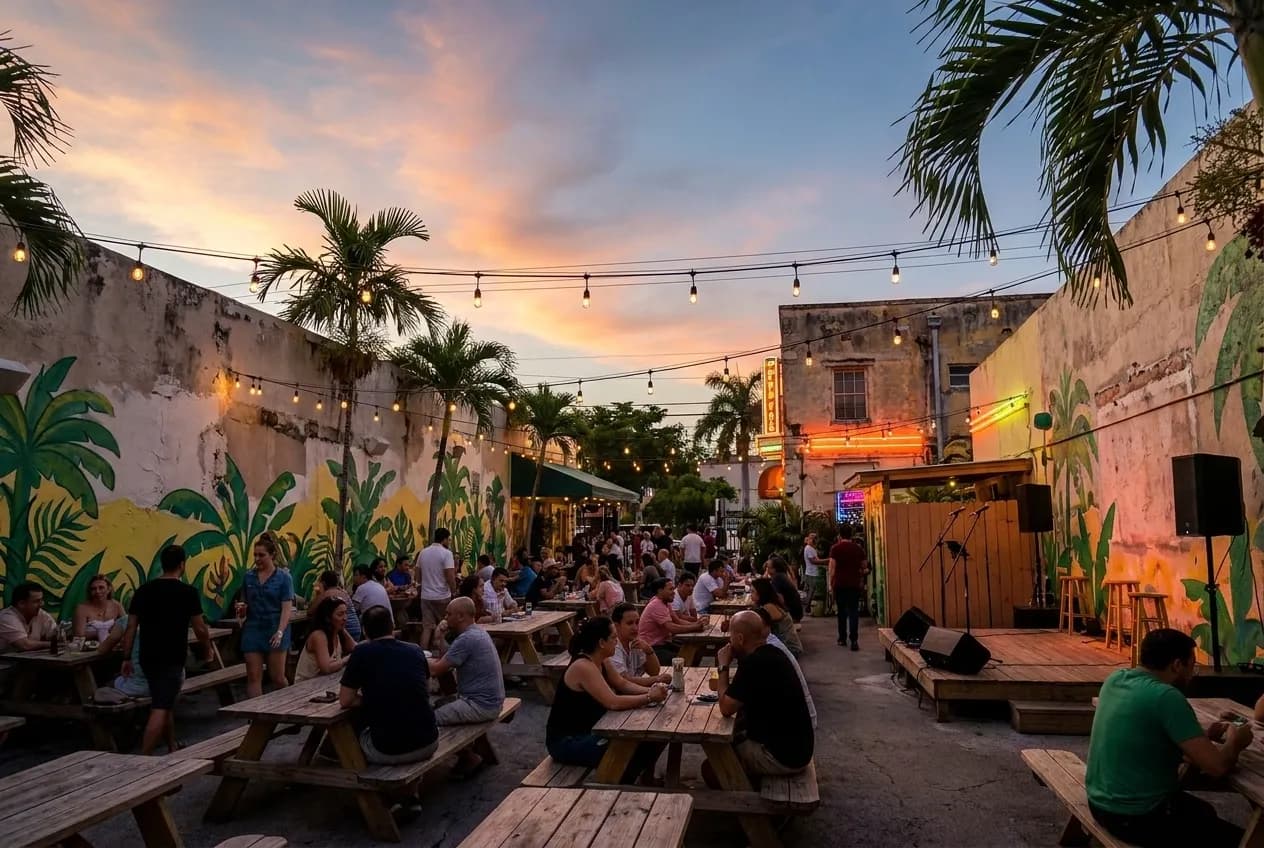 String lights and palm fronds frame an outdoor stage on Calle Ocho in Little Havana at dusk, with pastel storefronts glowing under warm evening light and concrete sidewalks still wet from afternoon rain.