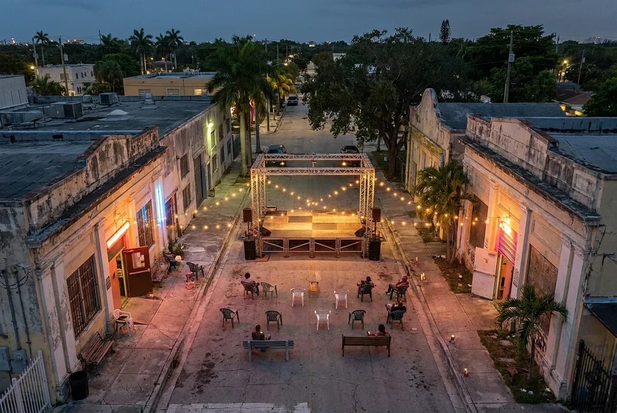 Open-air concert stage under colored lighting with metal railings and concrete floor in a Little Haiti performance yard surrounded by warehouse buildings at dusk.