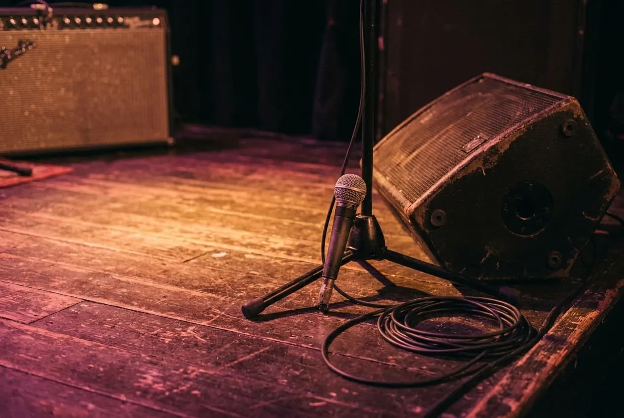 Close view of small concert stage with microphone stand, amplifiers, warm amber spotlights, and wooden floor in an intimate club setting.
