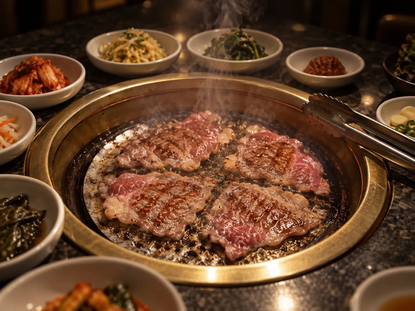 A smokeless tabletop grill set into a dark table with marbled beef slices cooking on the grate and small dishes of side vegetables ringed around it