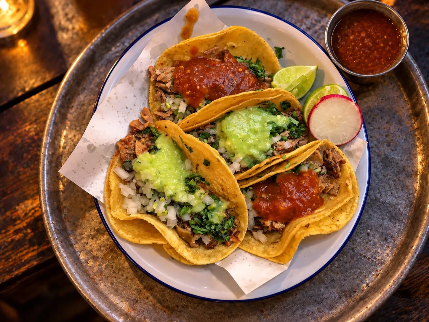 Four street tacos on freshly pressed corn tortillas with chopped onion, coriander, lime wedges and a bowl of salsa on a scratched metal tray
