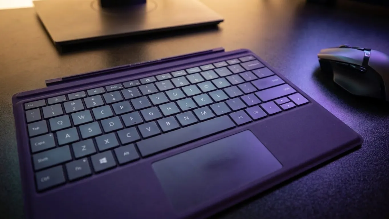 RGB mechanical keyboard and gaming mouse on desk surface with monitor displaying first-person shooter gameplay