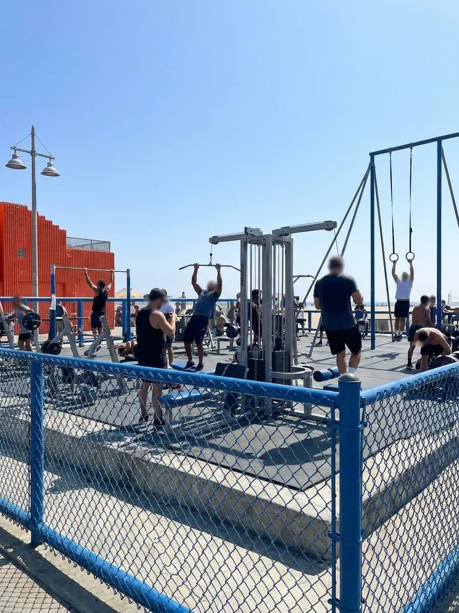 Muscle Beach Venice outdoor gym with chain-link fence, pull-up bars, weight platforms, and rings visible inside the gated area