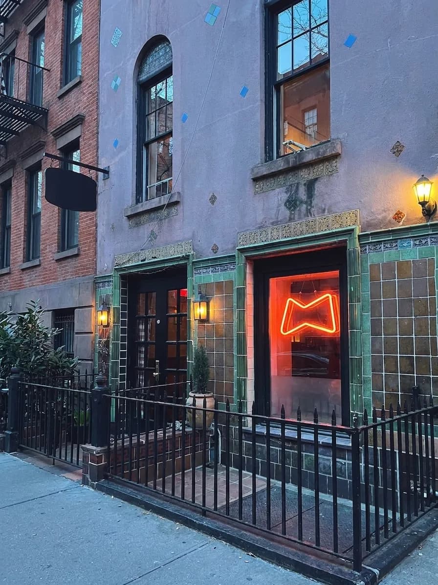 Night exterior of a green-tiled West Village pizza tavern with warm lantern light and a red neon glow in the window