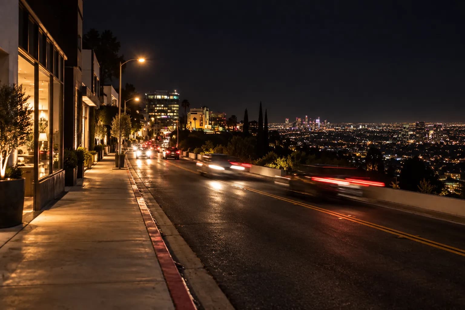 A ridge road at night with lit frontages on one side and the city grid glittering far below where the land drops away, headlights streaking