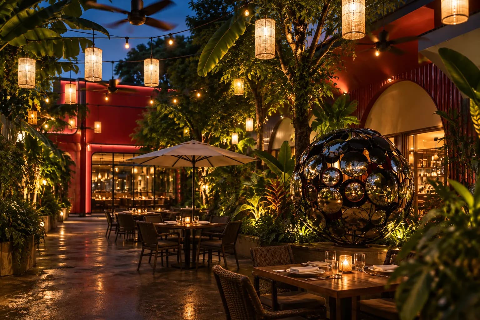 A planted restaurant courtyard just after sunset with tropical foliage against the walls, empty wooden tables, strung bulbs and ceiling fans