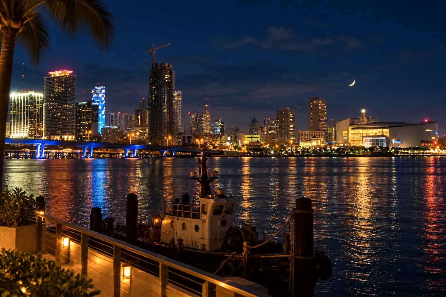 A working river at night with downtown towers lit and reflected in the water, a moored boat in the foreground and a warmly lit terrace opposite