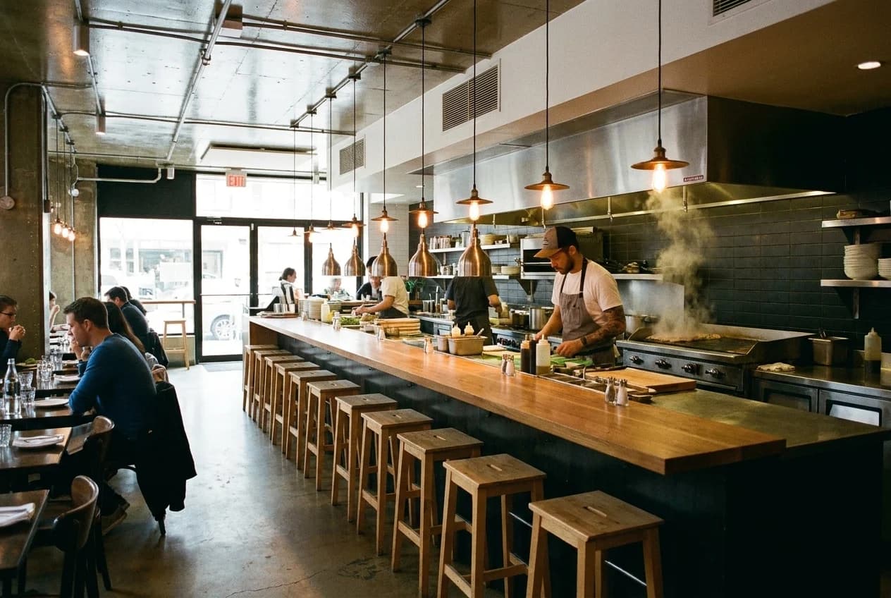 Empty counter stools facing an open kitchen with pendant lights and a cook at the grill in an East Village restaurant