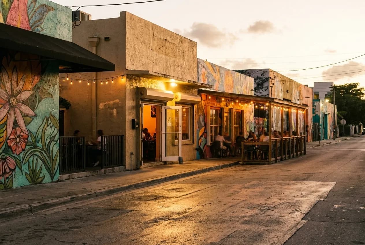Low-rise stucco buildings and murals along a quiet Wynwood side street at dusk, warm light spilling from restaurant windows onto the pavement.