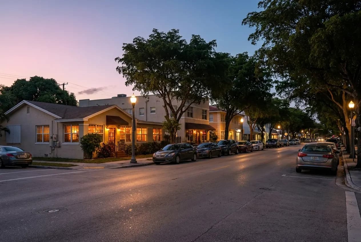 Coral Way at dusk with two-lane traffic, low apartment buildings, palm trees and warm storefront lights glowing along the sidewalk