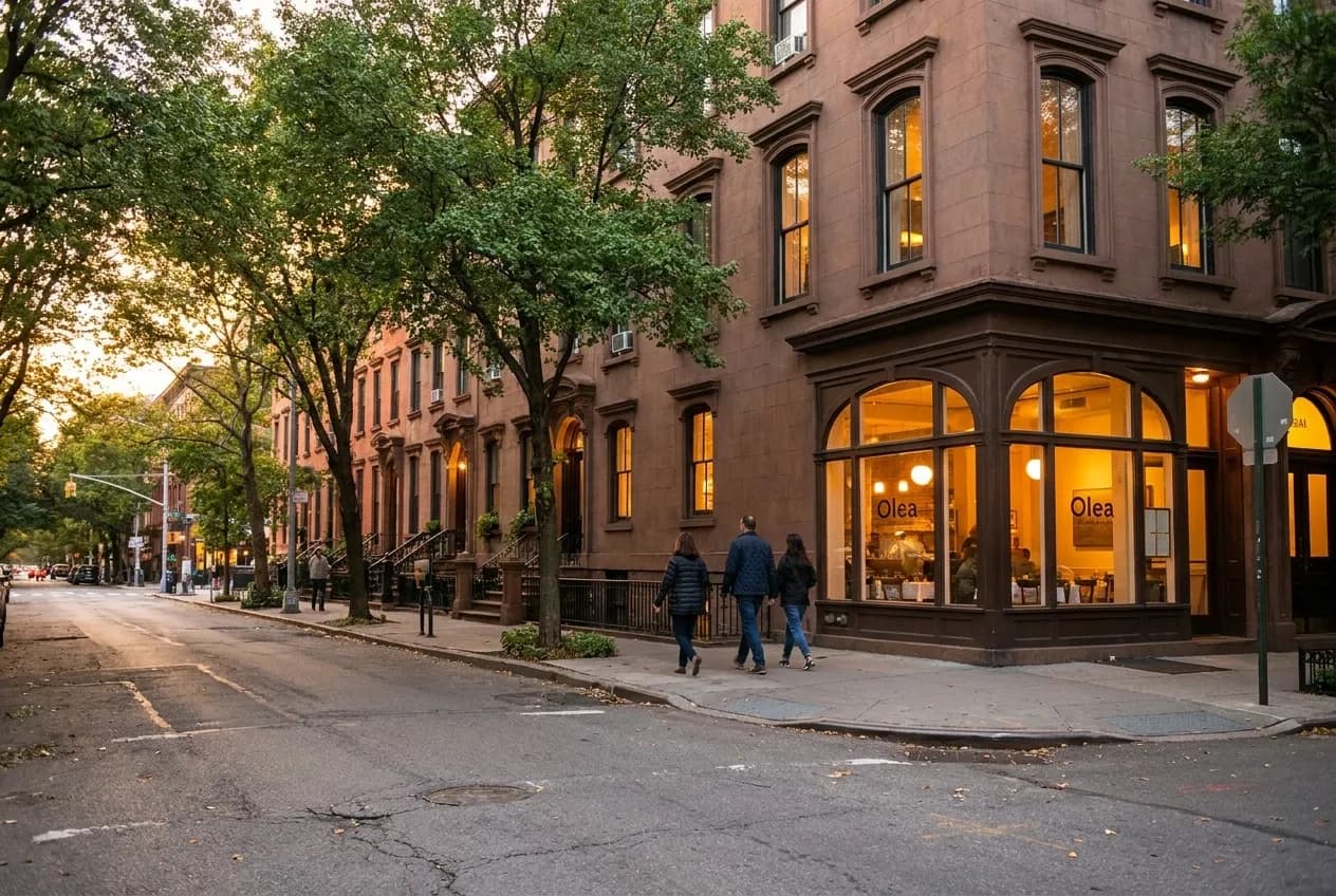Tree-lined Fort Greene avenue at dusk with brownstone stoops and soft bistro light spilling onto sidewalk near Brooklyn Academy of Music