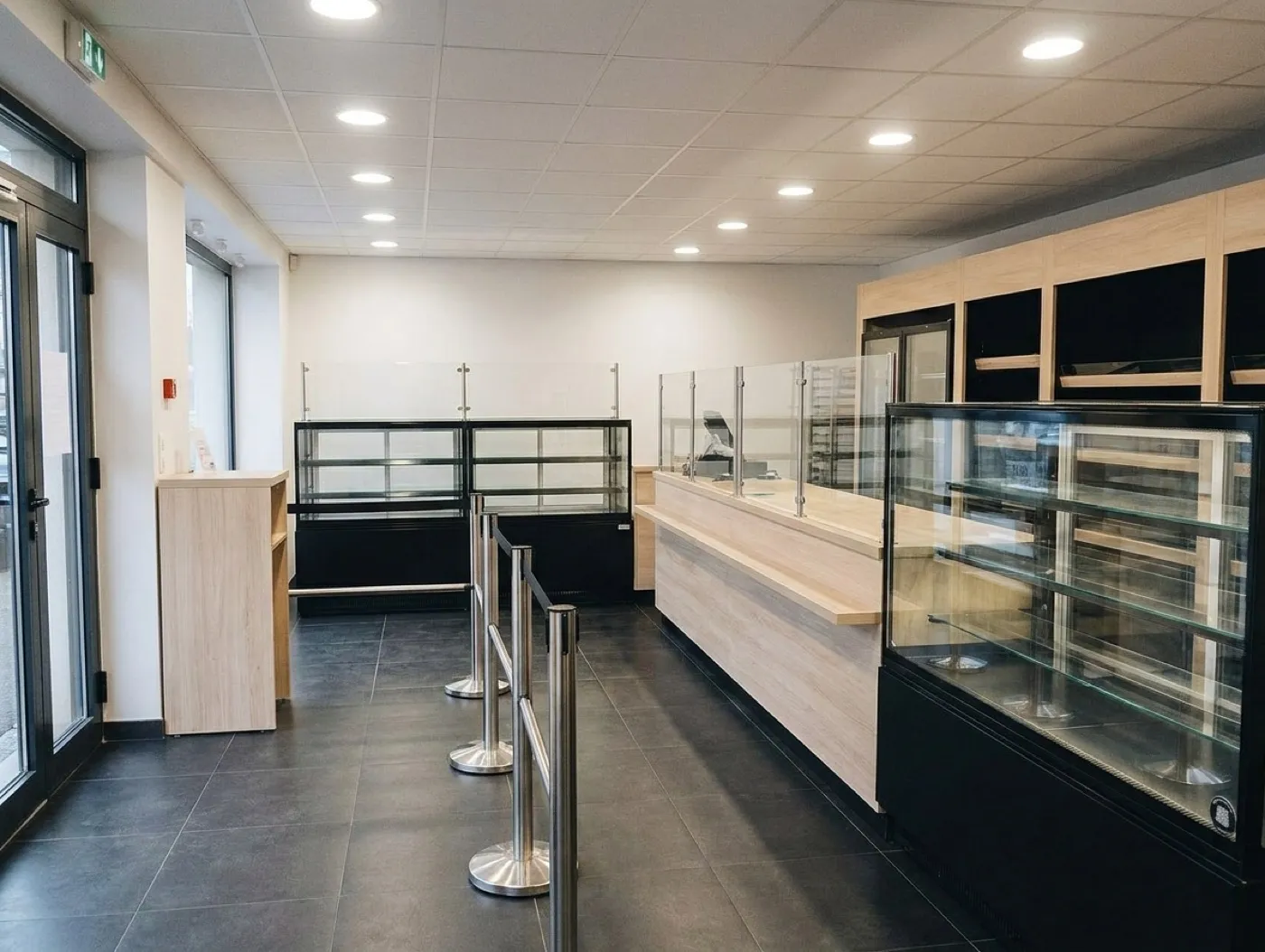 A clean modern retail interior with a pale wood counter, glass display cases and a queue rail, empty before opening