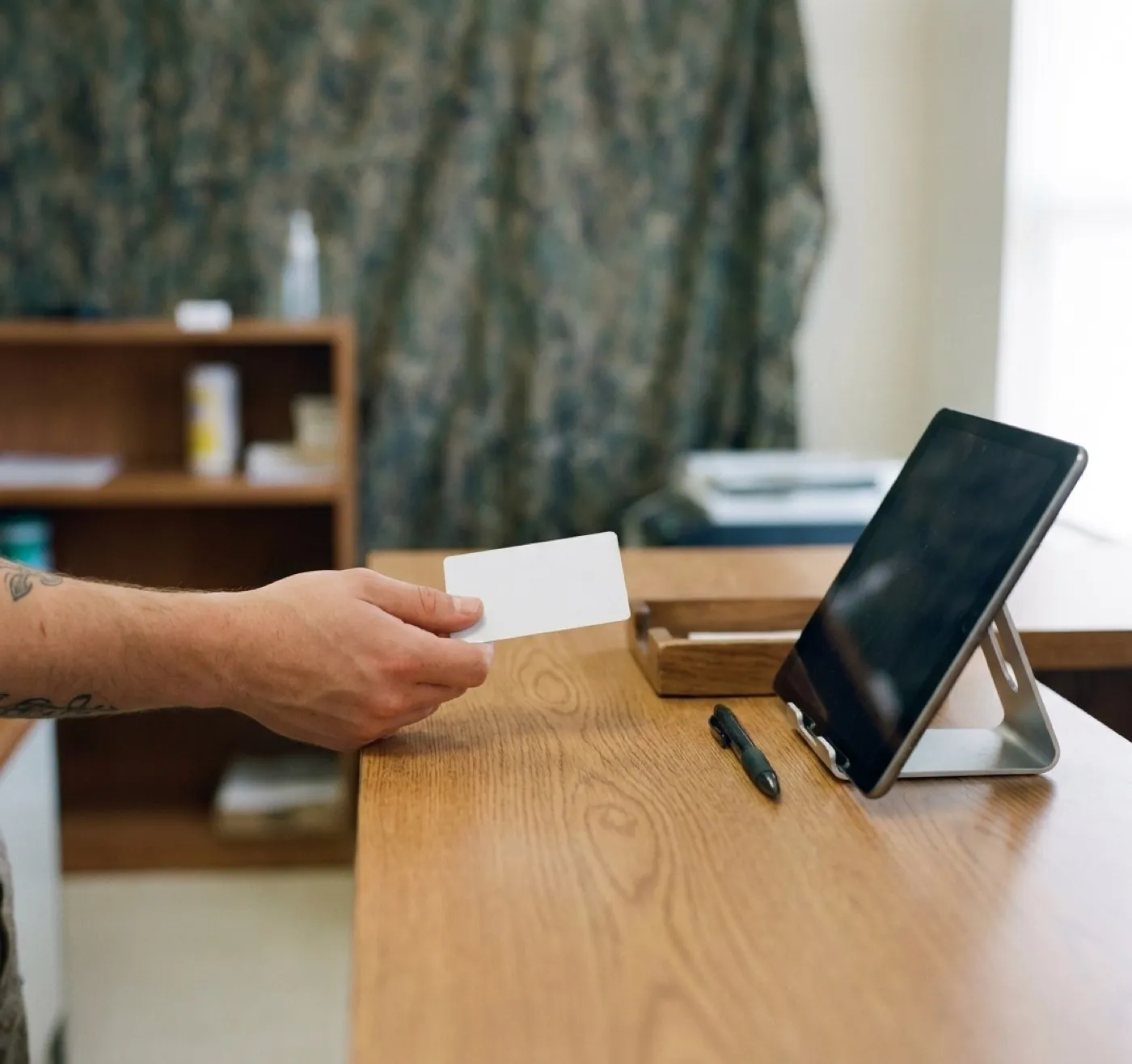 A hand holding a state identification card at a check-in desk with a tablet screen turned away from the camera