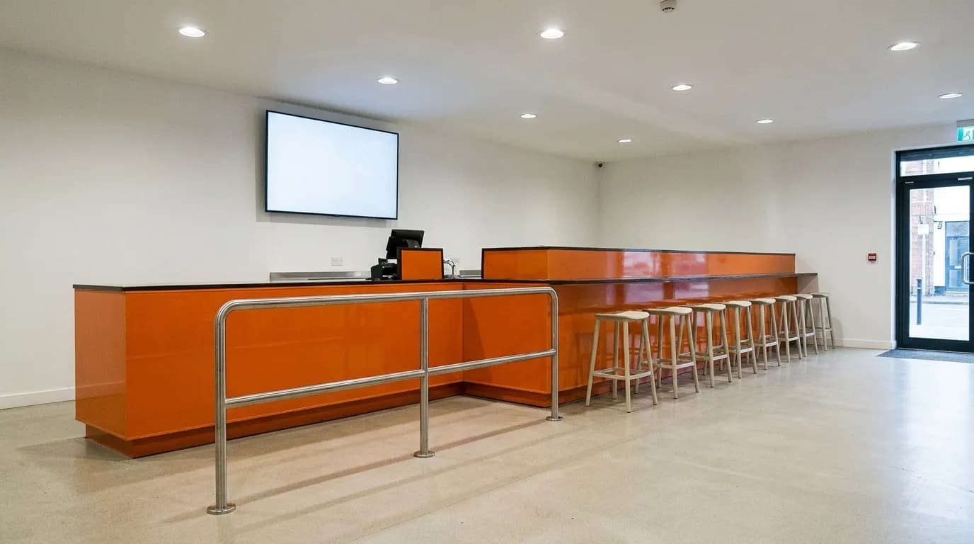 A clean retail counter with a queue rail, a screen mounted on the wall behind it and a row of empty stools, shot from the customer side of an empty room