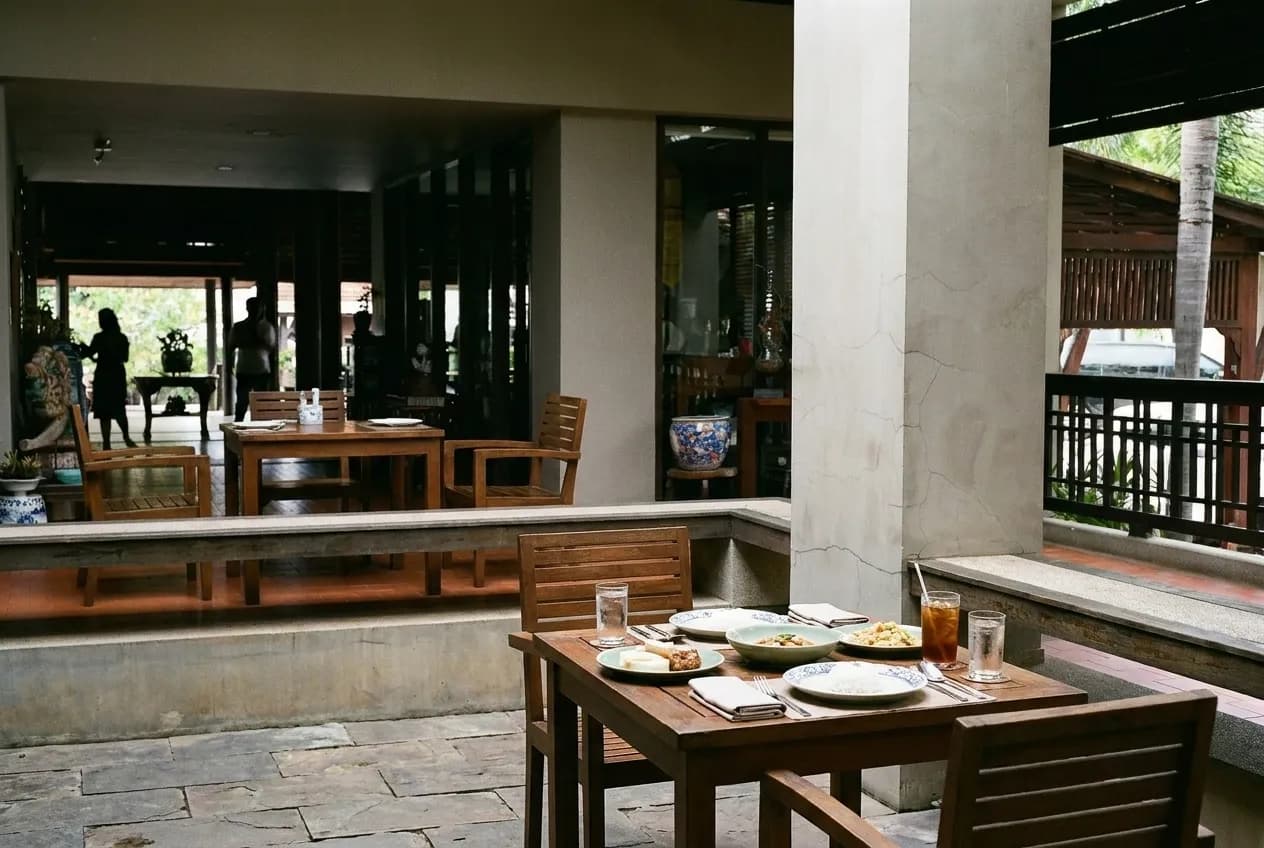 A shaded restaurant patio with wooden tables and a low wall onto the pavement