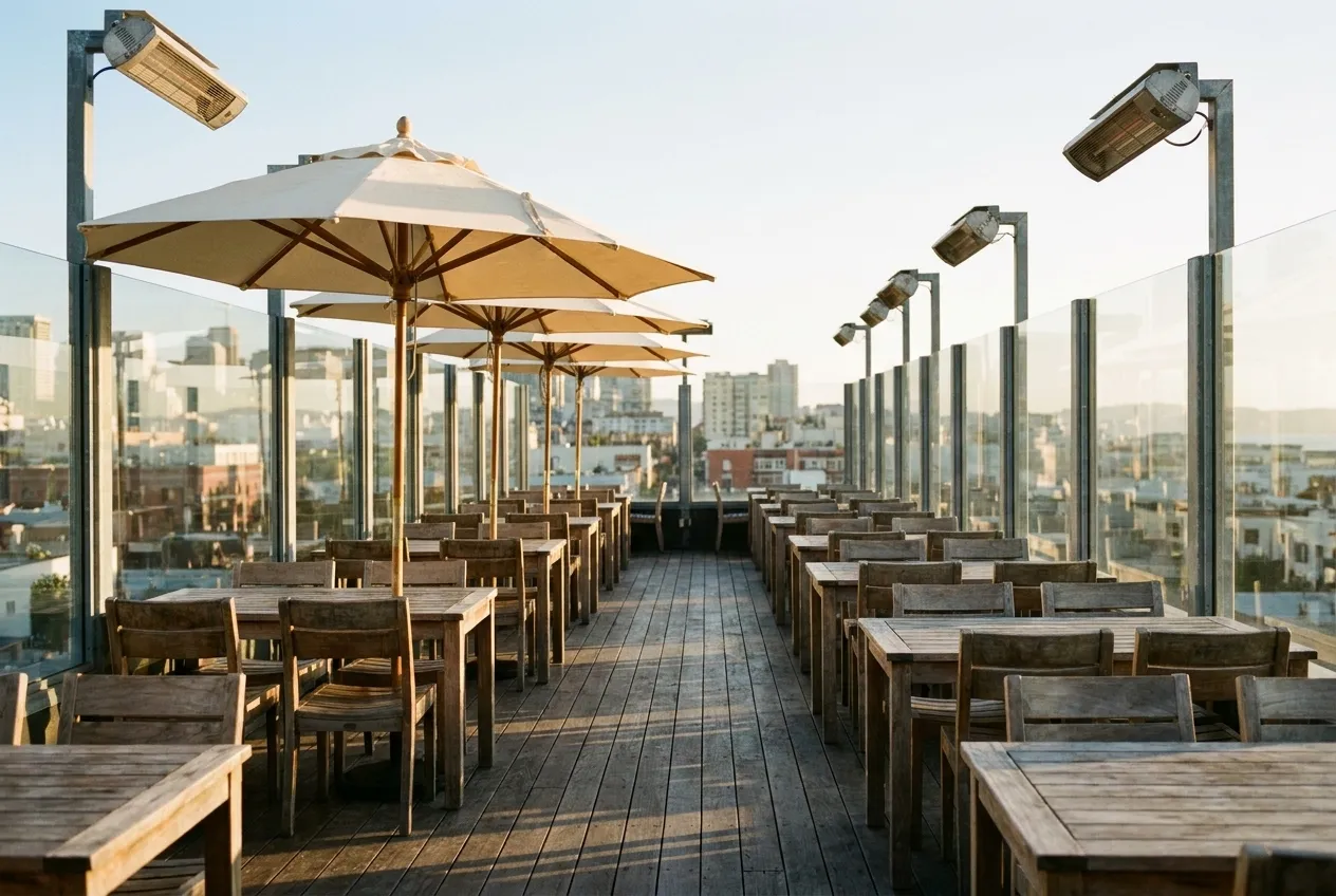 Wide view of an open-air rooftop bar with wooden tables, umbrellas, and glass wind barriers in San Francisco's Mission District at sunset.
