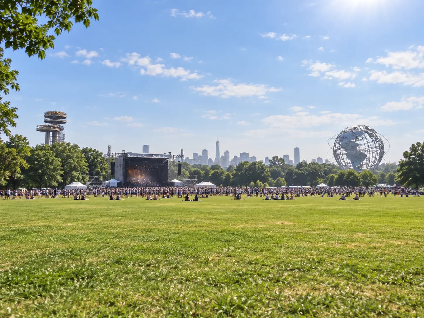 A large open park field with a festival stage in the distance and crowds under a summer sky
