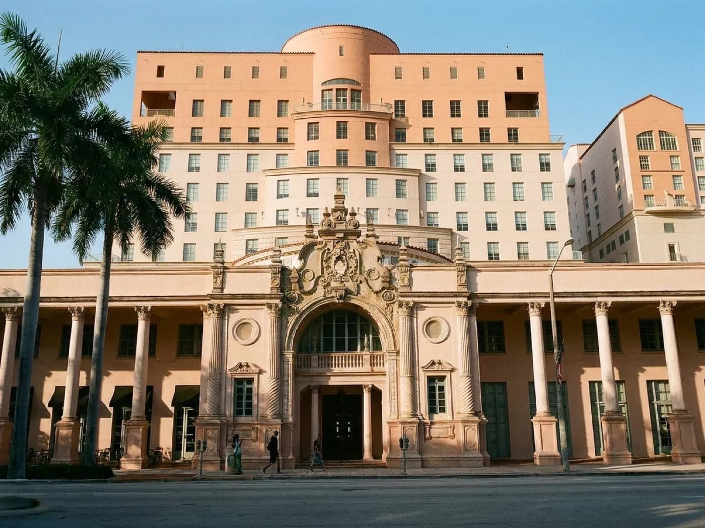 A Mediterranean revival building with arcaded walkways and barrel-tile roof on a wide palm-lined Coral Gables street in late afternoon
