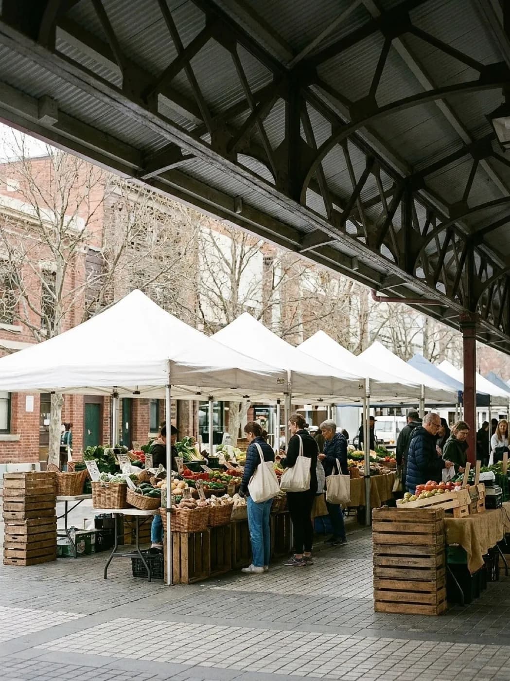 A busy outdoor greenmarket in a city square with produce stalls under white canopies and shoppers with canvas bags