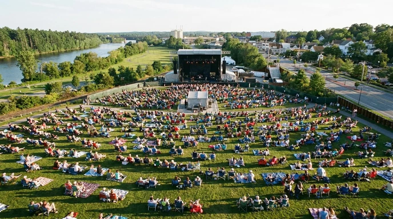 A crowd of picnic blankets on grass in front of a distant open-air stage under trees