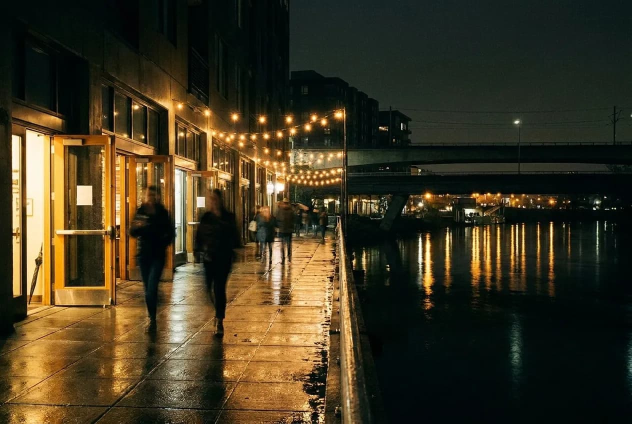 A lit sidewalk at night with warm light spilling from open doorways and figures blurred in motion