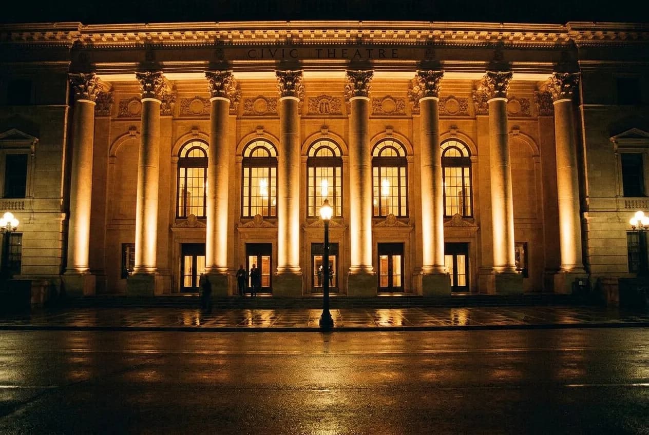 A colonnaded civic theatre facade lit from below at night with glowing arched windows and wet pavement