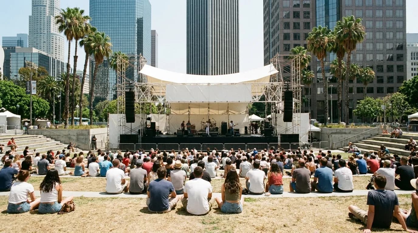 An outdoor lunchtime concert in a downtown Los Angeles park, people sitting on grass and low steps in front of a small stage with palm trees and office towers behind