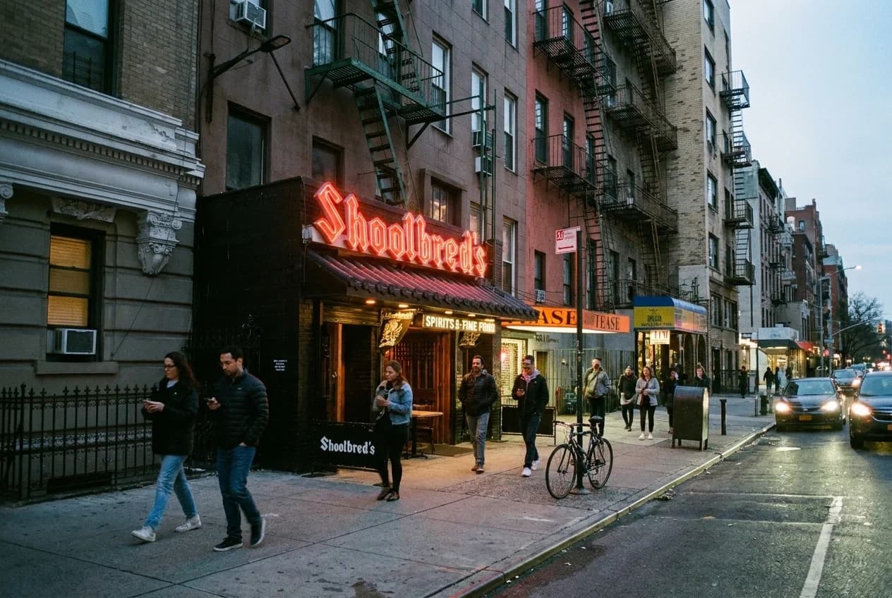 Avenue A at dusk with neon bar signs and pedestrians walking past East Village storefronts