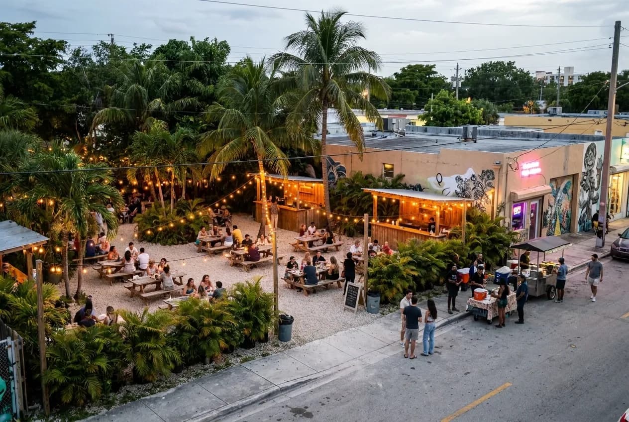 A gravel path winds through dense tropical plantings and picnic tables under string lights in an open-air Miami garden venue at twilight, with wooden bar structures visible in the background.