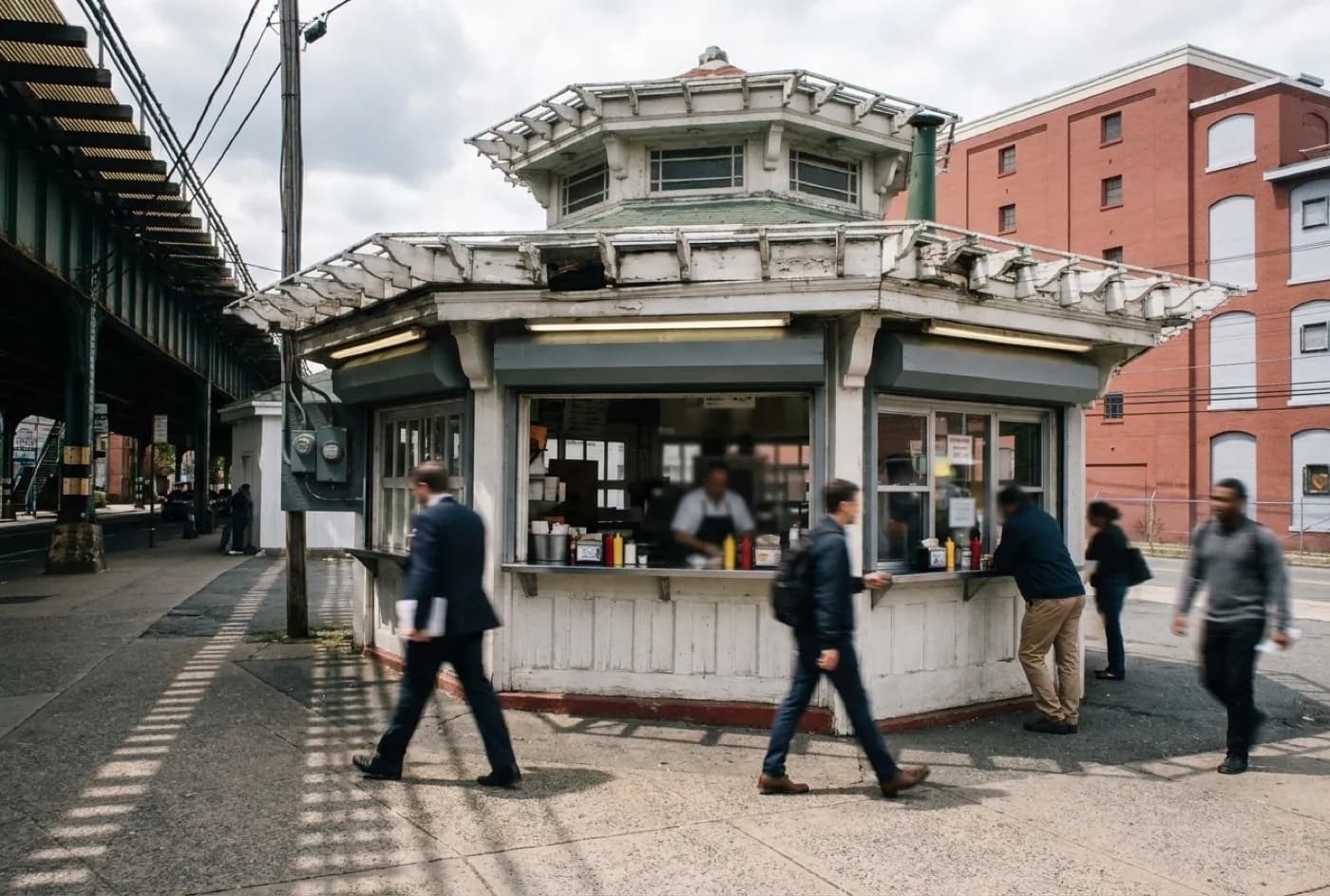 A corner hot dog counter under a subway overpass in New York with a narrow standing ledge and commuters passing on the pavement
