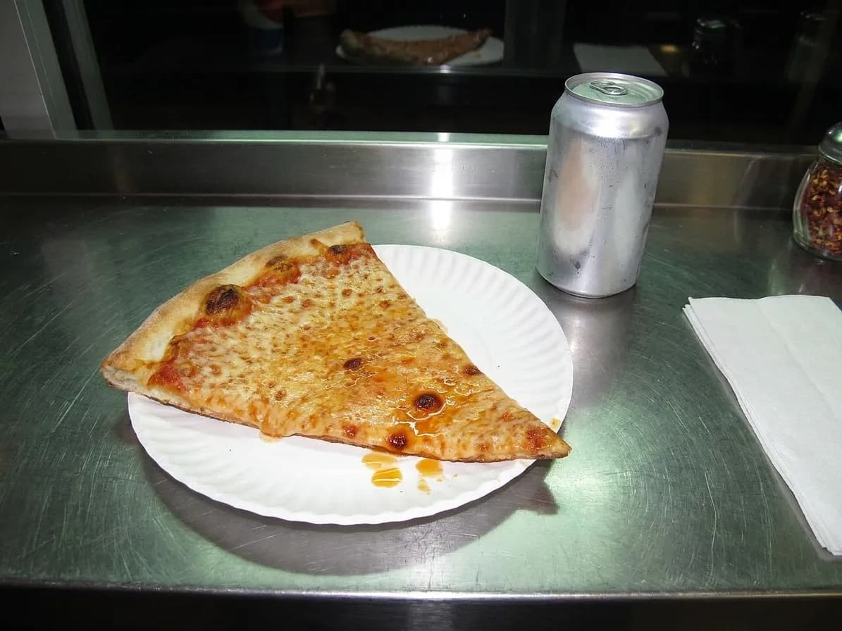 A plain cheese slice on a paper plate beside an unlabelled soda can on a scratched stainless counter under flat late-night fluorescent light