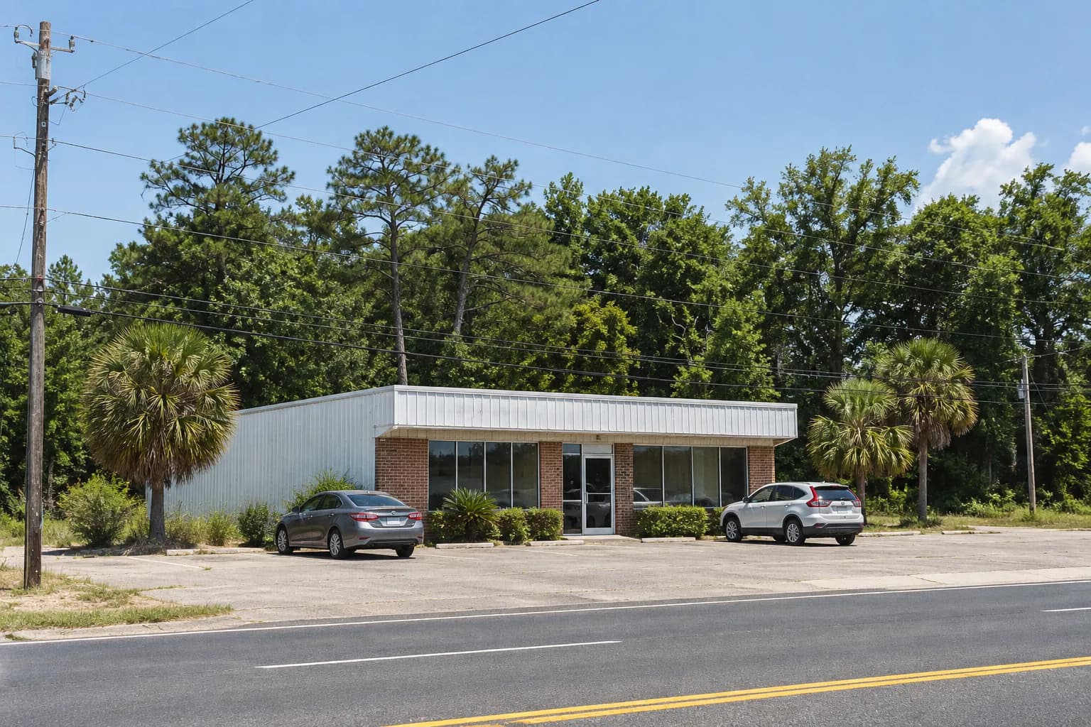 A very small single-unit roadside building across an empty parking apron in hard midday sun, cars parked at angles and short palm shadows