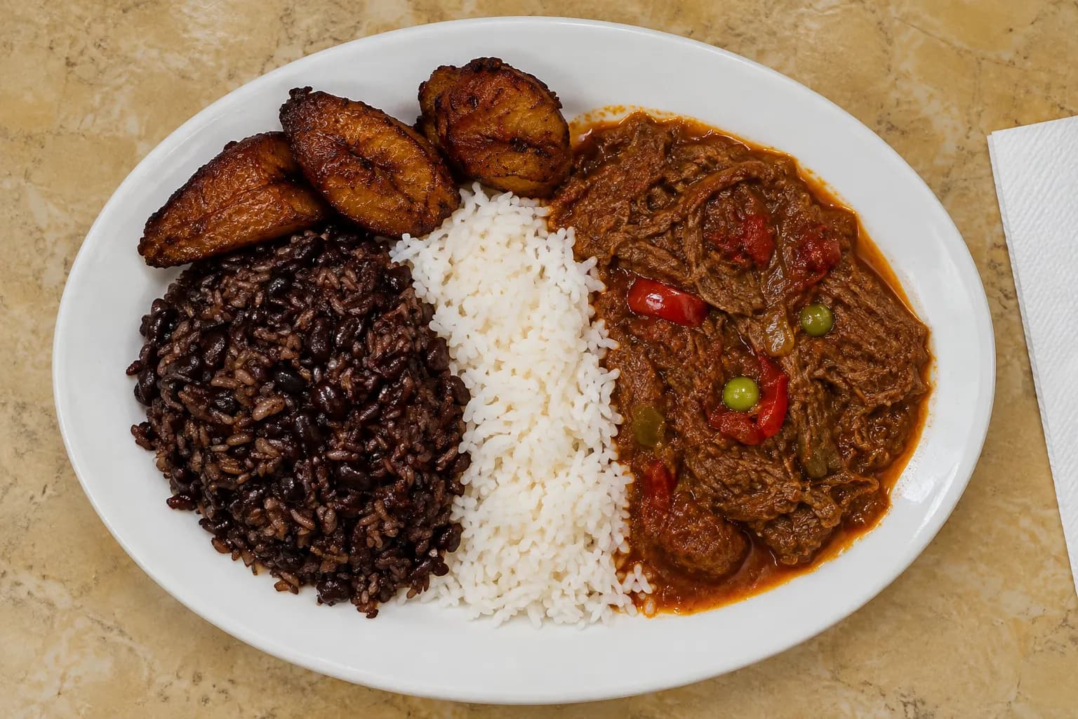 An oval plate of shredded stewed beef in red-brown sauce with white rice, black beans and caramelised fried plantains on a formica table