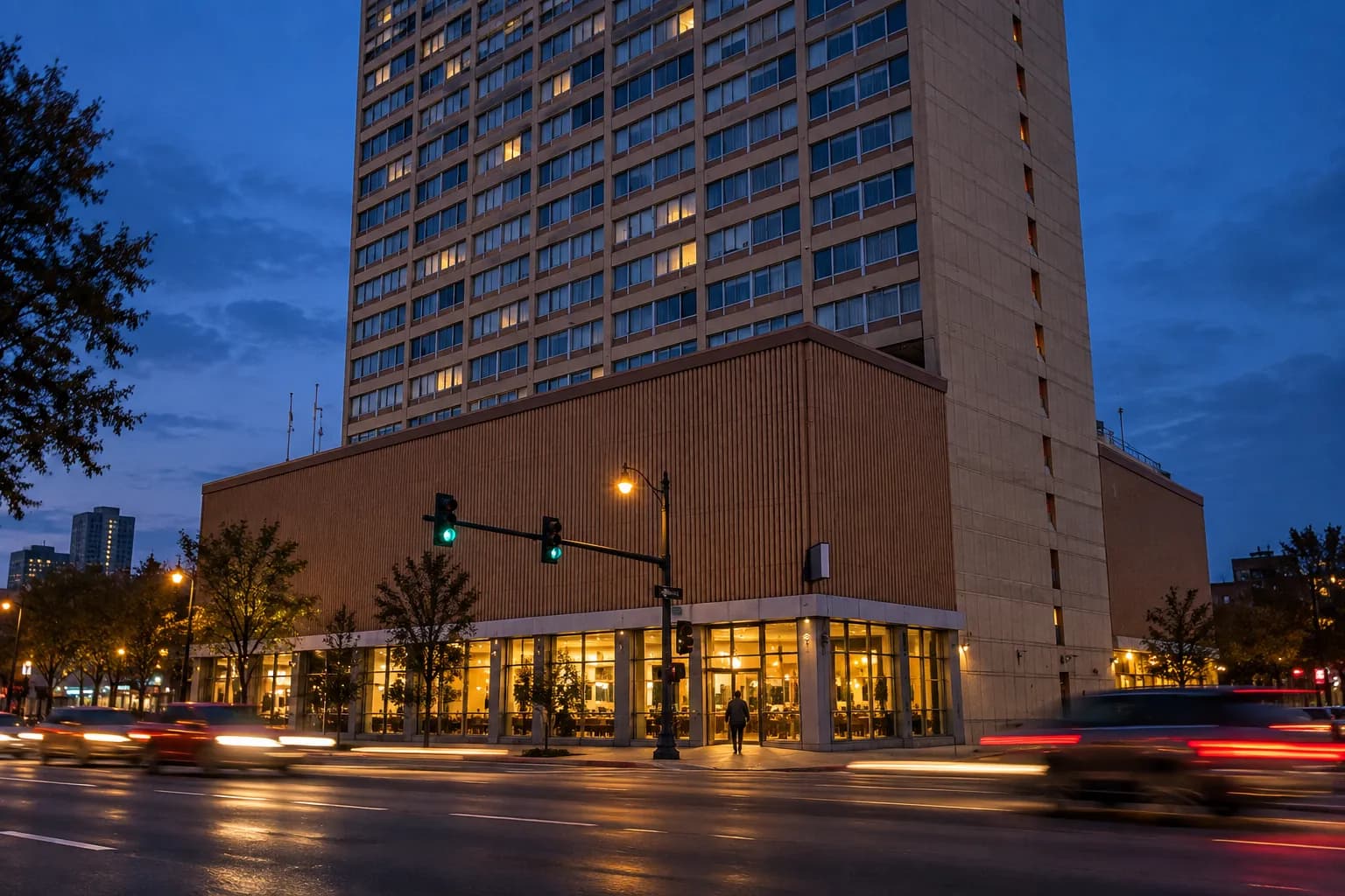 A painted corner building across a wide multi-lane road at blue hour, its ground floor warmly lit and traffic streaking past in the foreground