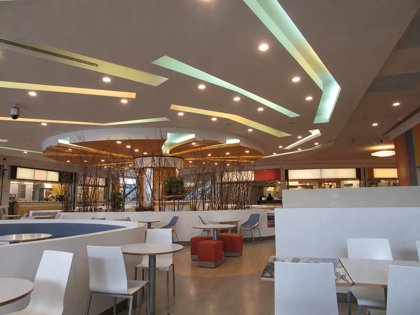 Indoor food court on a lower shopping level with rows of tables and moulded chairs, perimeter counters and a curving escalator beyond