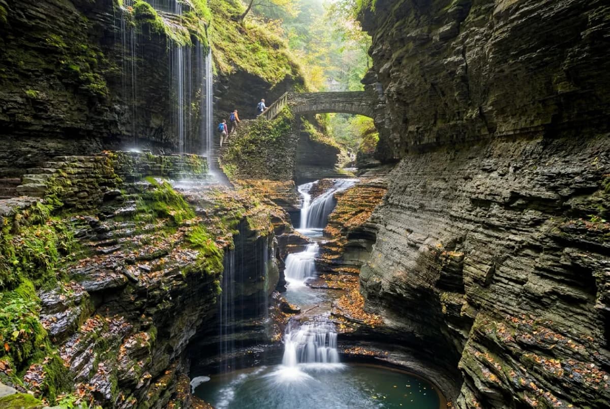A dramatic slate gorge with waterfalls and a stone footbridge at Watkins Glen State Park near Seneca Lake, with hikers for scale