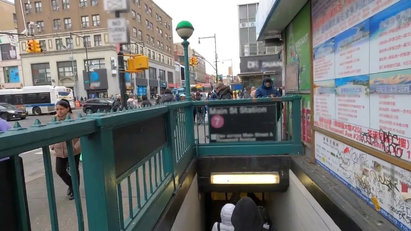 Green-railed subway entrance at a crowded Flushing street corner in Queens with poster-covered walls and winter foot traffic