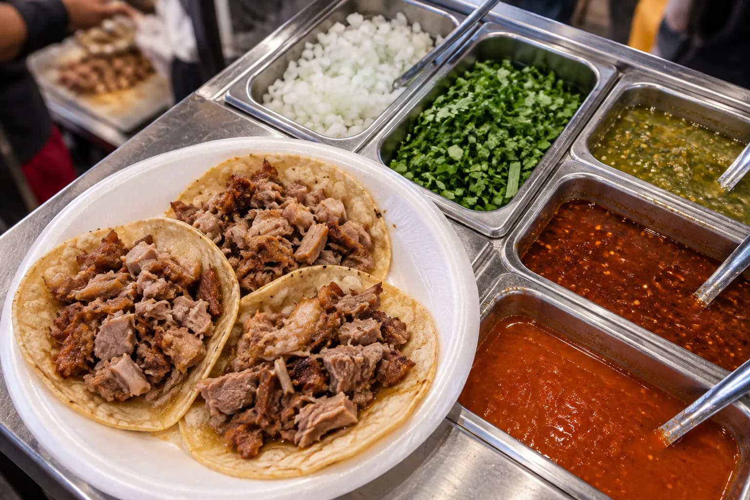 A tray of carnitas tacos on a styrofoam plate beside a self-serve condiment bar of chopped onion, coriander and salsas under market lighting